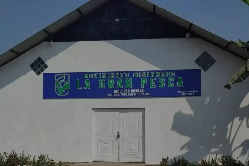 Imagen de un templo de la iglesia Movimiento Misionero La Gran Pesca, ubicado en San Antonio, ciudad de San Juan, Pueblo Viejo. El edificio tiene una pared blanca con un globo que dice 'Movimiento Misionero La Gran Pesca'. La puerta es blanca y está rodeada de piedras blancas y rosas.